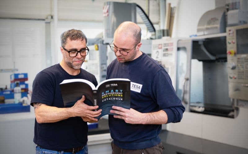 Two men in a workshop reading a catalog titled "Smart Instrument Solutions." Machinery and industrial equipment in the background.